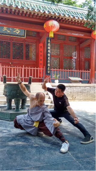 A monk in gray robes and a man in black practice low martial arts stances in a traditional temple courtyard.